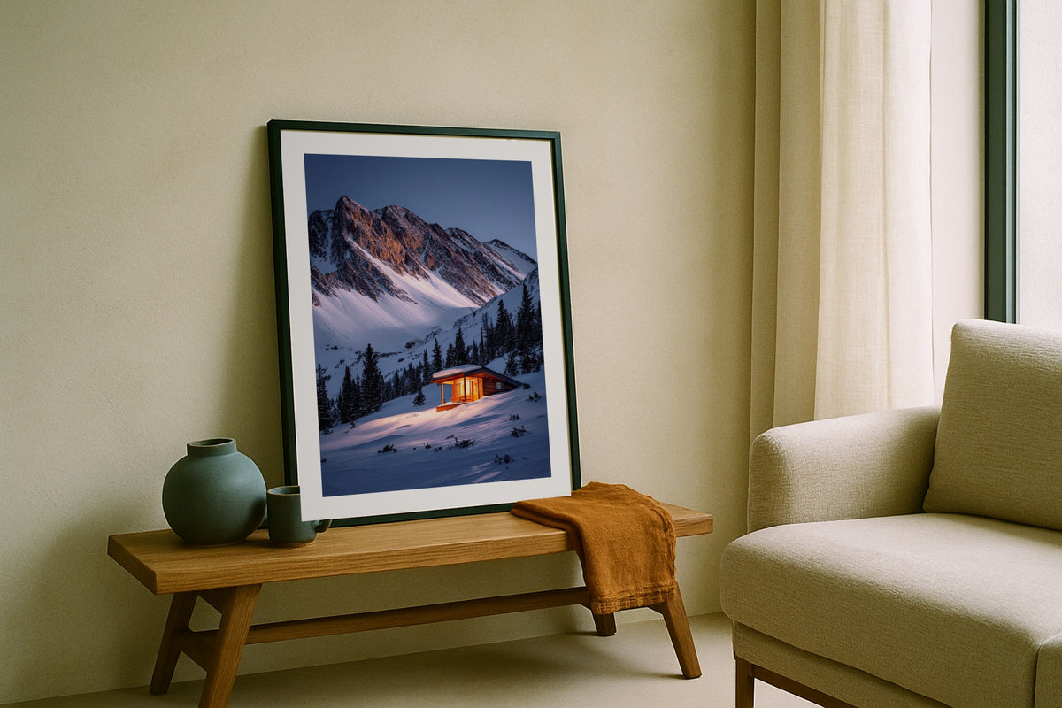 Mountain Cabin in Winter wall art print, framed with a white border, in a leaning on bench